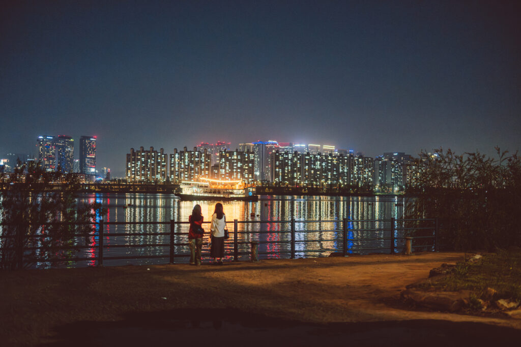 Two people standing beside the Han River with a brightly lit Seoul skyline reflected in the water