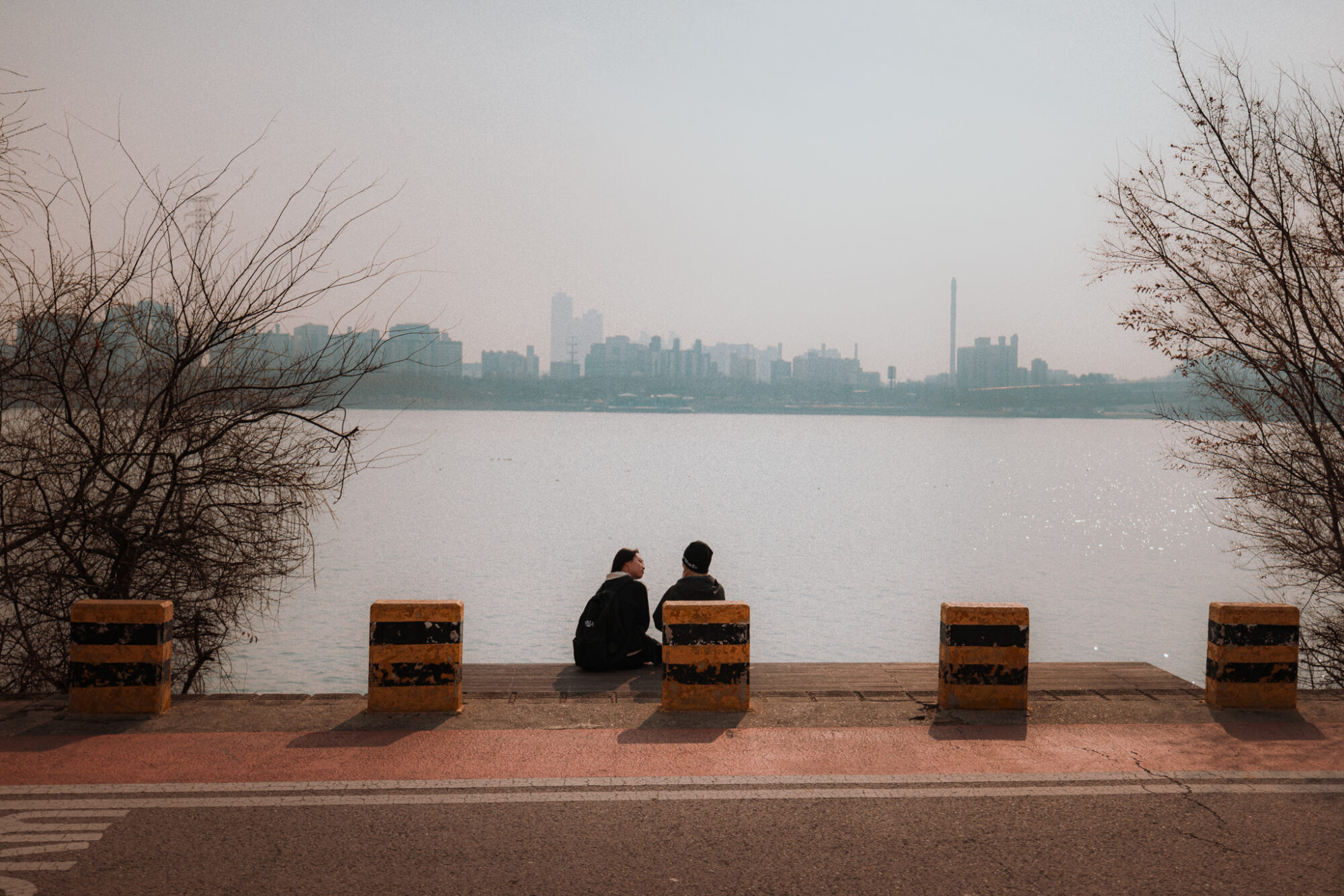Two people sitting beside the Han River with Seoul's skyline visible through the haze