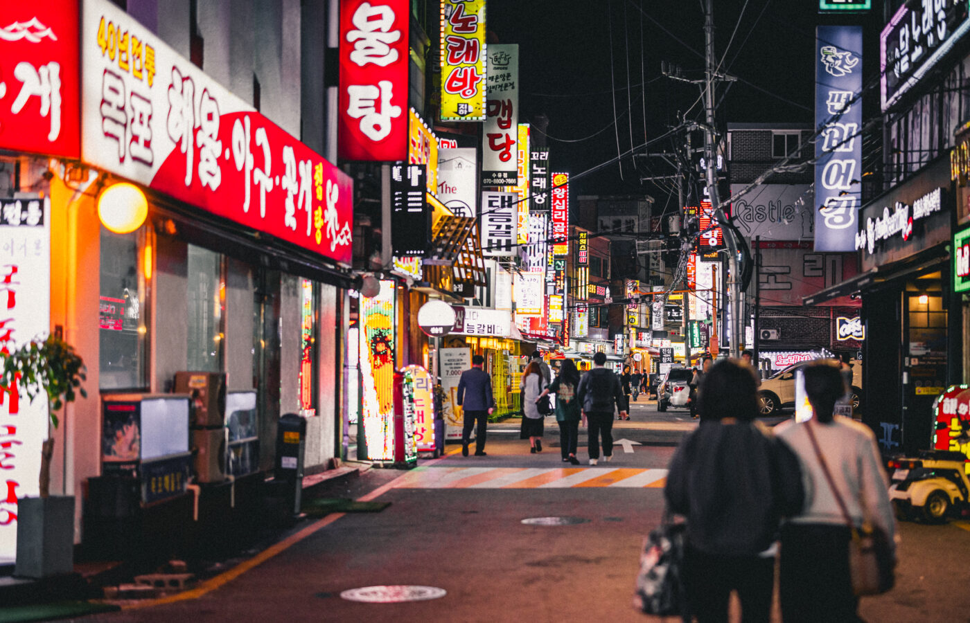 Pedestrians walking through a busy Seoul nightlife street lined with bright restaurant, bar and karaoke signs