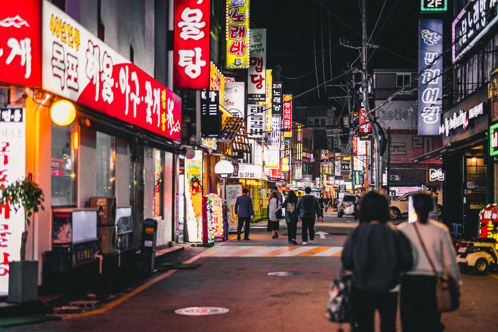 Pedestrians walking through a busy Seoul nightlife street lined with bright restaurant, bar and karaoke signs