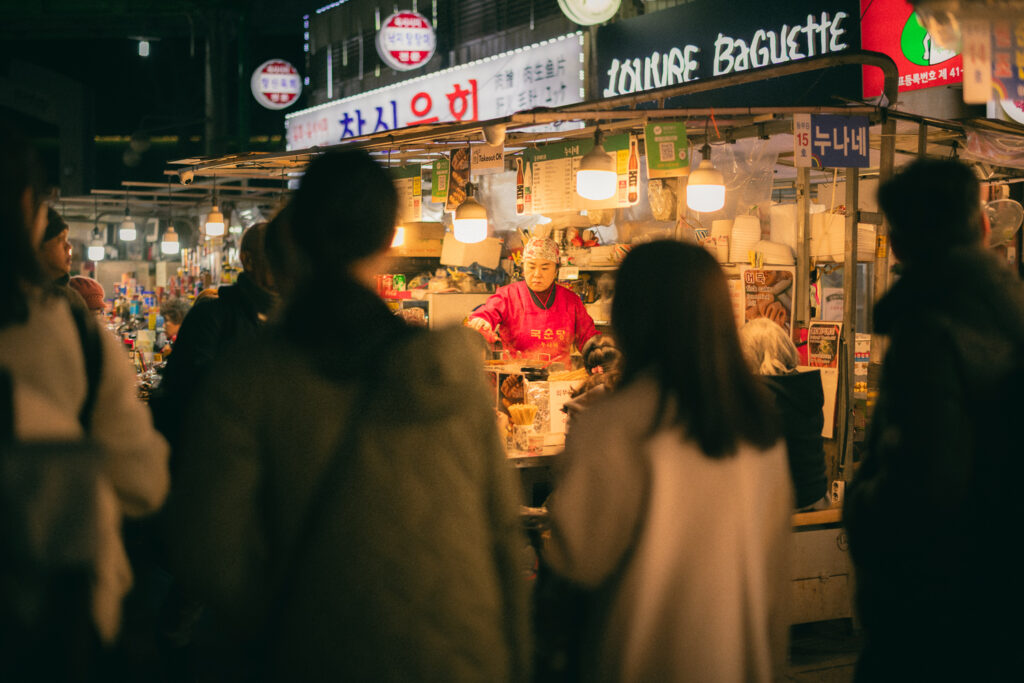 Street food vendor serving customers at a brightly lit Korean market stall at night