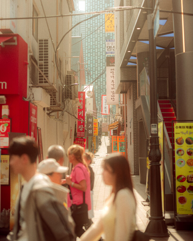 Pedestrians passing a narrow Seoul alley lined with Korean restaurant signs in warm afternoon light