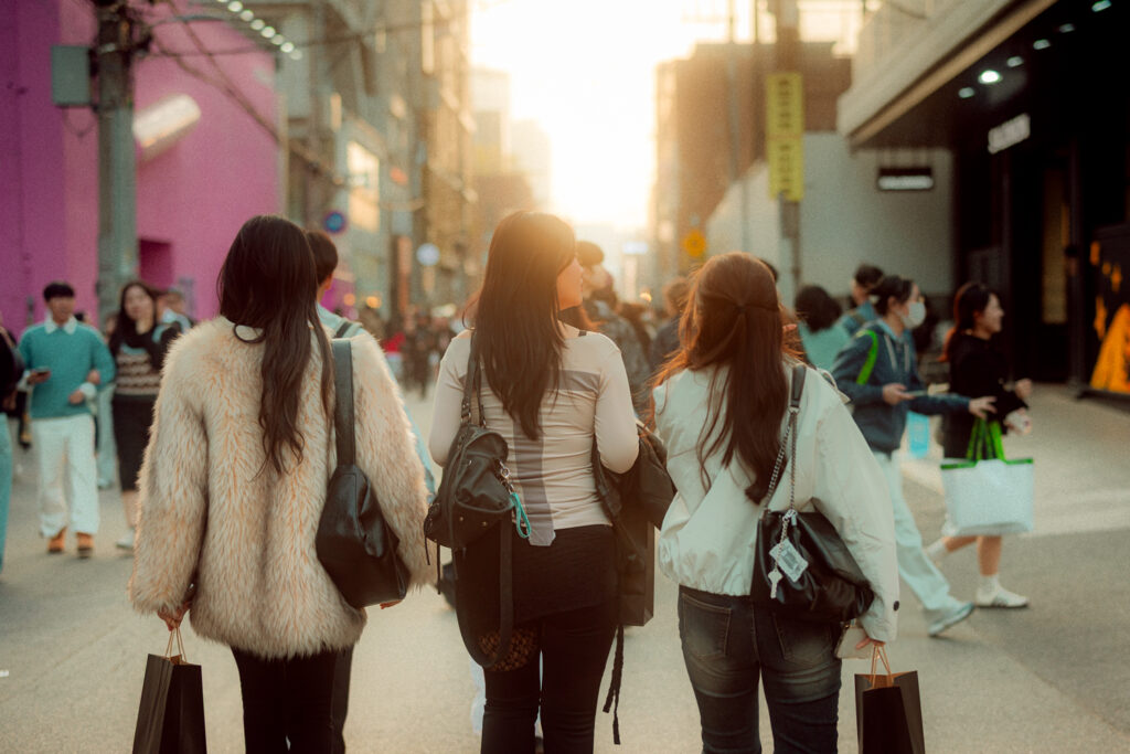 Three women carrying shopping bags through a busy Seoul street in warm evening sunlight
