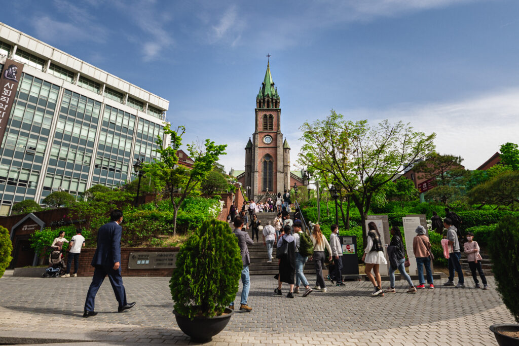 Visitors walking up the steps toward Myeongdong Cathedral, surrounded by green trees on a sunny day in Seoul