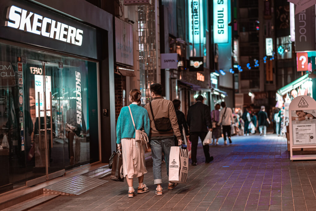 Two shoppers carrying bags past a Skechers store on a brightly lit shopping street in Myeongdong, Seoul