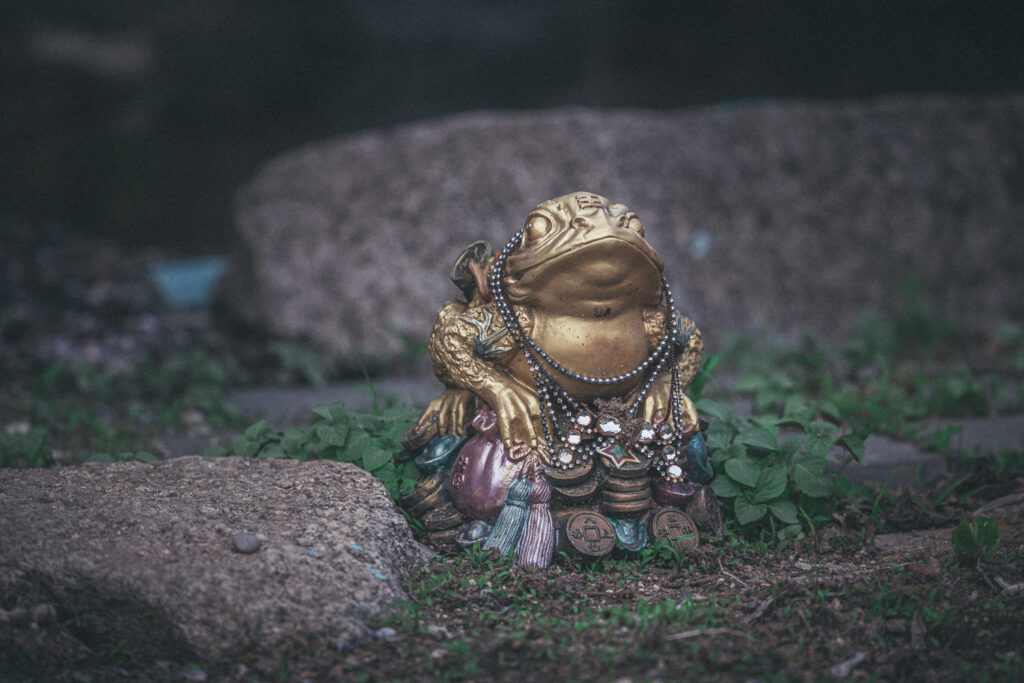 Golden frog figurine draped in bead necklaces sitting among stones and low plants.