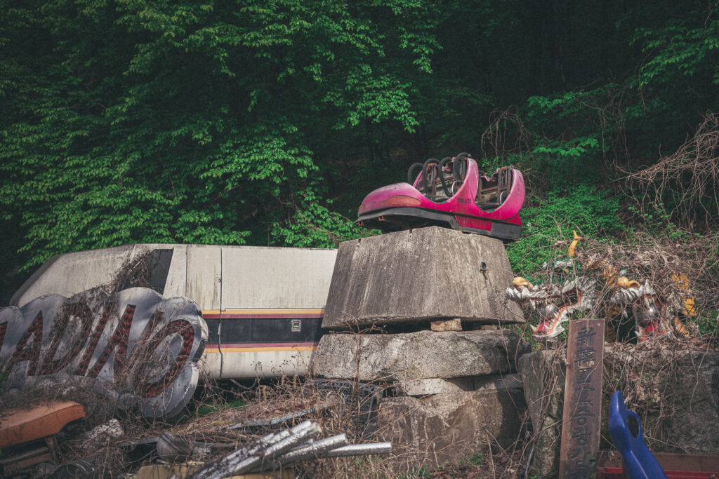 Pink amusement ride car perched above concrete barriers and discarded equipment beside dense greenery.