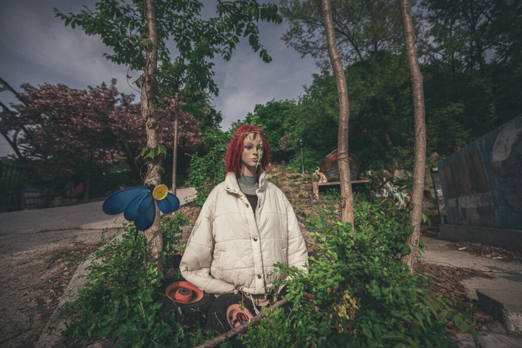 Red-haired mannequin in a white padded jacket standing among plants and trees at Yongma Land.
