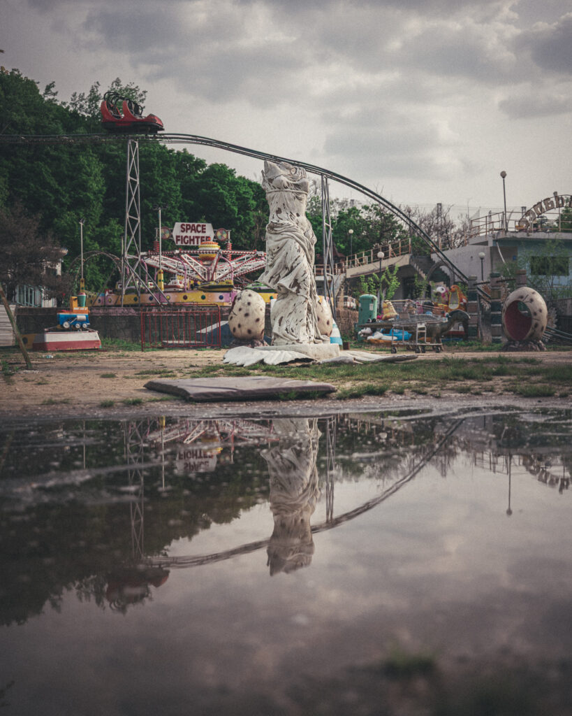 White statue and elevated ride track reflected in a large puddle beneath a cloudy sky at Yongma Land.