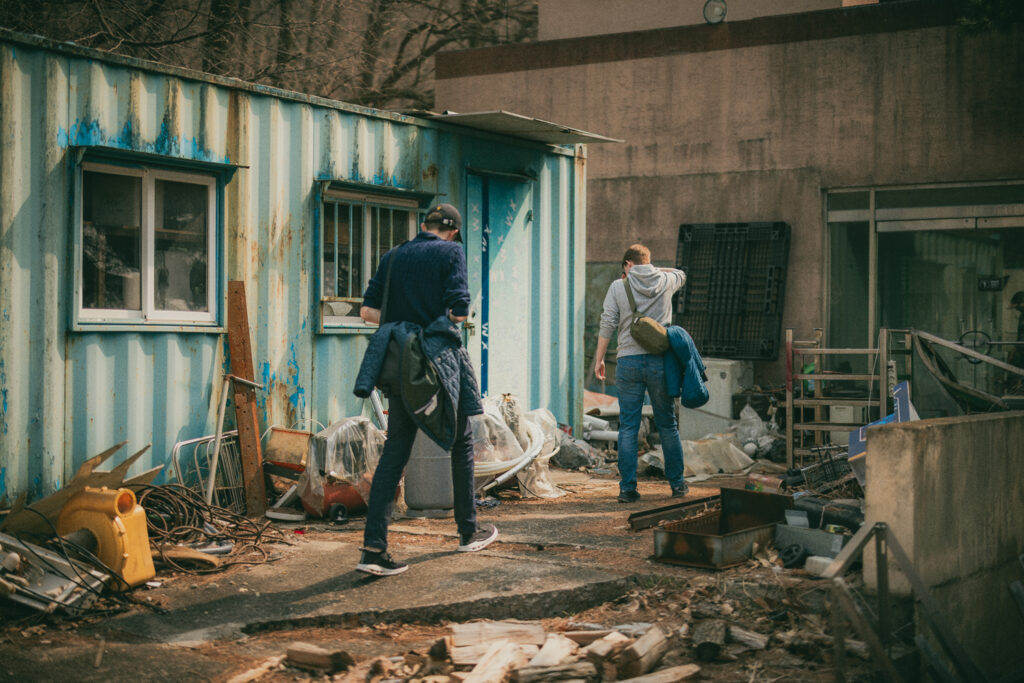 Two visitors walking past scattered equipment and a weathered blue and white building at Yongma Land.